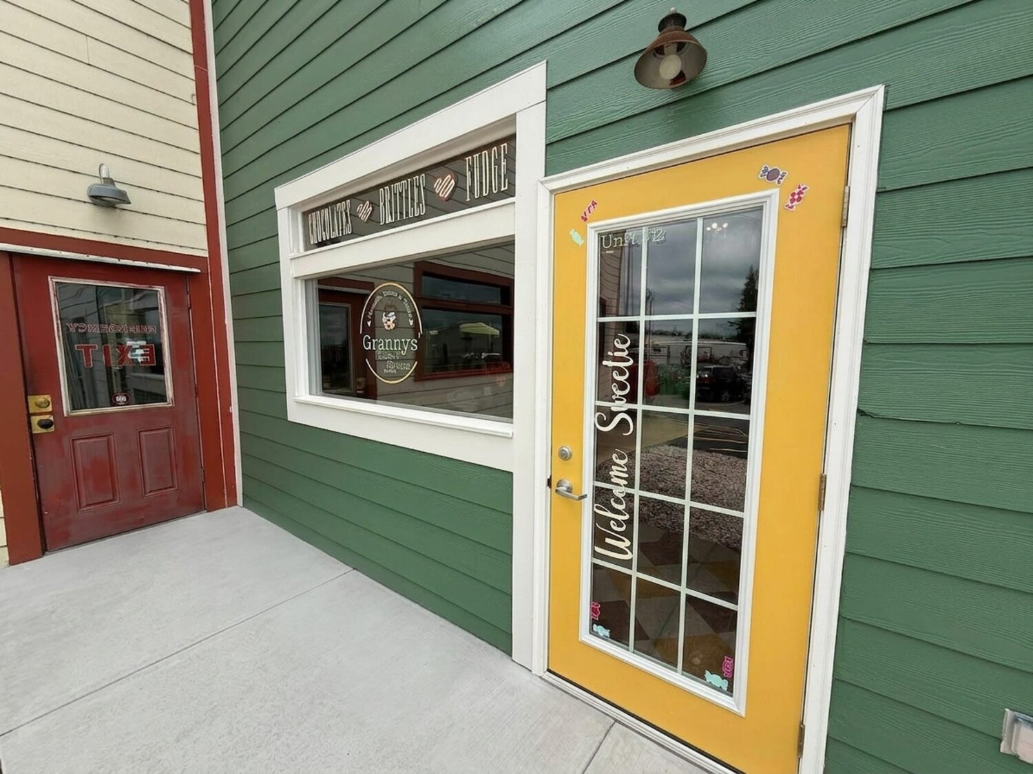 The storefront of Granny's House of Sweets: green siding, a marigold 'Welcome Sweetie' door, and a display window lettered Chocolates, Brittles, Fudge with the Granny's logo.